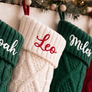 Può includere: Quattro calze natalizie in maglia appese in fila. Due sono verde scuro con i nomi Noah e Miles ricamati in bianco, una è color crema con il nome Leo ricamato in rosso e una è rossa con il nome Owen ricamato in rosso.
