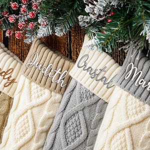 Peut inclure: Quatre bas de Noël en tricot torsadé beige, crème, gris et taupe, chacun avec un nom brodé sur le revers : Sandy, Judith, Crastal et Martha. Les bas sont présentés avec des branches de conifères et des baies rouges.