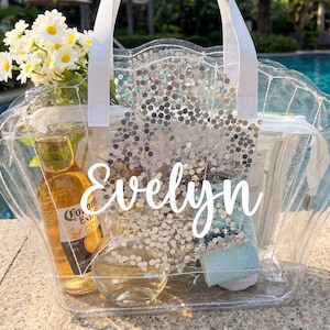 May include: A clear, scalloped-edge tote bag with white handles, filled with items including a bottle of beer, flowers, and a small bottle. The name "Evelyn" is written in white script on the front. The bag is set near a pool.