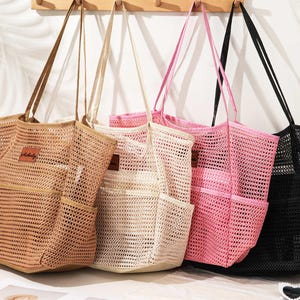 May include: Four mesh tote bags in shades of tan, cream, pink, and black. Each bag has a matching handle and a front pocket. A small brown label is attached to the front of each bag. The bags are hanging on wooden pegs.