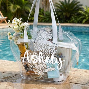 Könnte beinhalten: Eine transparente Tragetasche mit Gegenständen wie einer Bierflasche, einem Buch und Blumen. Die Tasche hat weiße Griffe und den Namen "Ashely" in weißer Schrift. Die Tasche steht an einem Pool.