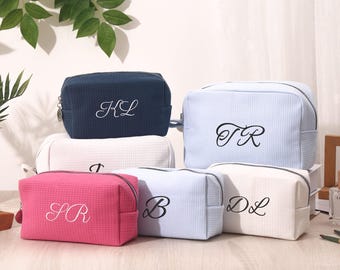 May include: A collection of personalized cosmetic bags in various colors, including navy blue, pink, and light blue. Each bag features embroidered initials and a zippered closure. The bags have a textured, waffle-like pattern.