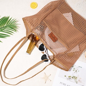 Pu&ograve; includere: Una borsa tote in rete marrone con manici lunghi, contenente una bottiglia, occhiali da sole e una piccola pochette. Una fetta di limone, una stella marina e una rivista sono nelle vicinanze. La borsa ha una piccola etichetta in pelle.