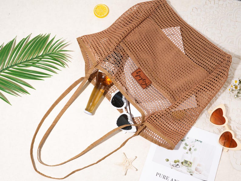 Pu&ograve; includere: Una borsa tote in rete marrone con manici lunghi, contenente una bottiglia, occhiali da sole e una piccola pochette. Una fetta di limone, una stella marina e una rivista sono nelle vicinanze. La borsa ha una piccola etichetta in pelle.