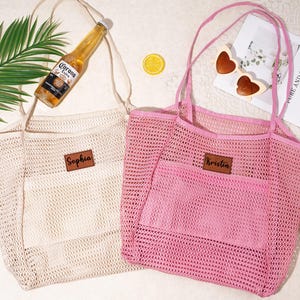 Pu&ograve; includere: Due borse a rete, una beige e una rosa, con manici abbinati e una tasca frontale. Ogni borsa ha una toppa in similpelle marrone con un nome sopra. Una bottiglia di birra, occhiali da sole e una rivista sono nelle vicinanze.