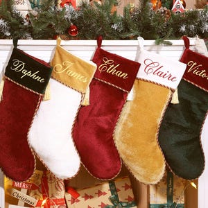 May include: Five personalized Christmas stockings in red, white, gold, and green velvet, each with a name embroidered in a complementary color. Gold tassels and decorative trim add detail. Festive holiday decor.