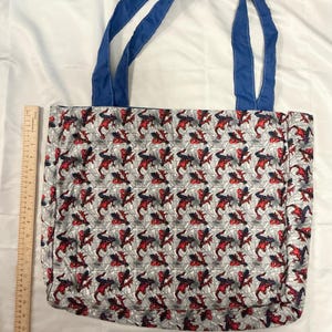 May include: A tote bag featuring a repeating pattern of a red, black, and blue superhero on a gray web background. The bag has blue handles and is made of fabric. The bag is approximately 15 inches wide.