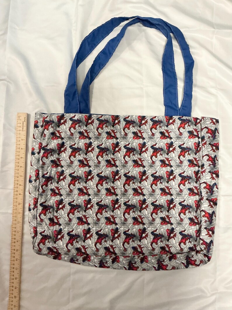 May include: A tote bag featuring a repeating pattern of a red, black, and blue superhero on a gray web background. The bag has blue handles and is made of fabric. The bag is approximately 15 inches wide.