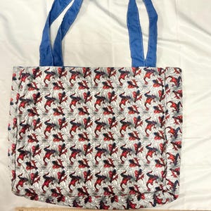 May include: A rectangular tote bag with blue handles. The bag's body features a repeating pattern of a red and black superhero figure on a gray background with a web design. The handles are a solid blue color.
