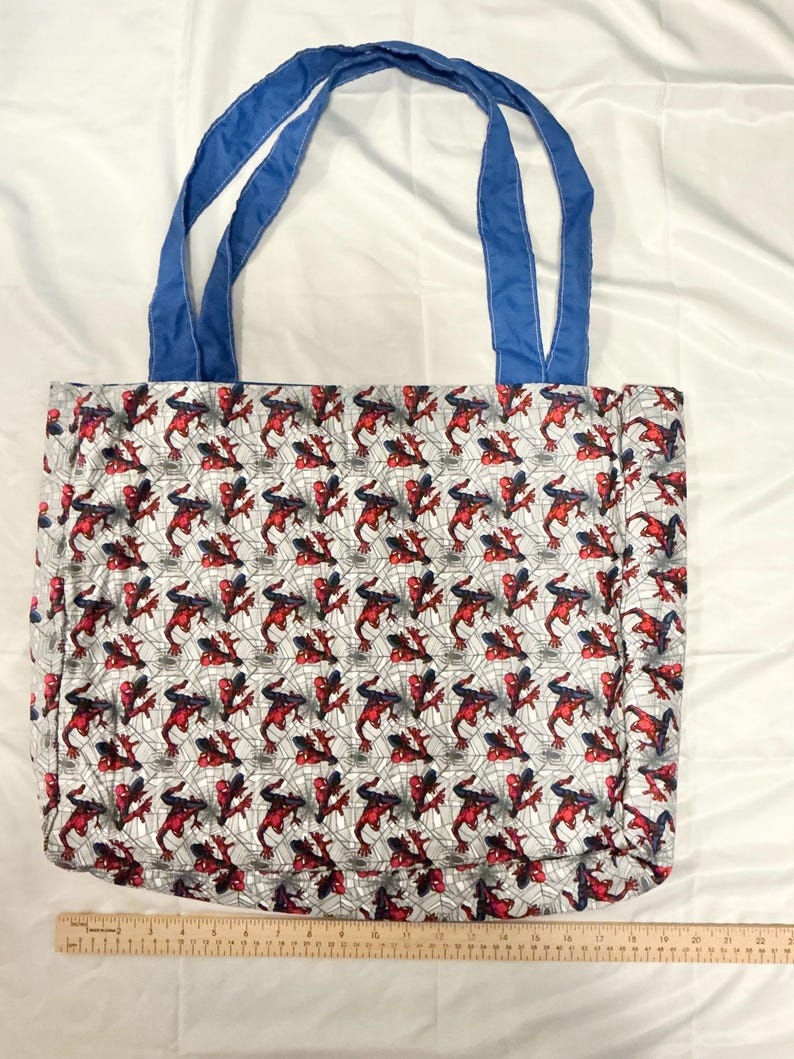 May include: A rectangular tote bag with blue handles. The bag's body features a repeating pattern of a red and black superhero figure on a gray background with a web design. The handles are a solid blue color.