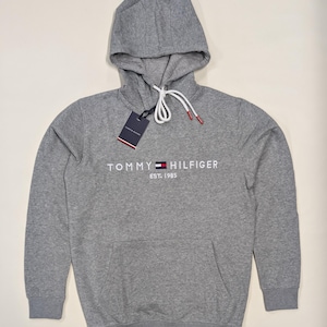 May include: A grey hooded sweatshirt featuring the Tommy Hilfiger logo in white. The sweatshirt has a drawstring hood, a front pocket, and a tag. The text "EST. 1985" is below the logo. A classic, casual garment.