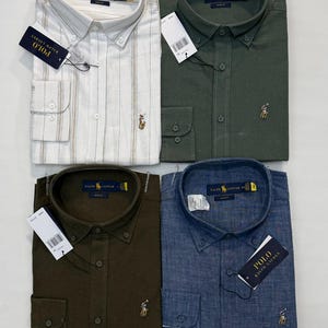 May include: Four folded button-down shirts in different colours: white with vertical stripes, dark green, olive green, and blue. Each shirt has a button-down collar, long sleeves, and a small embroidered logo. Each shirt has a tag that says "POLO RALPH LAUREN".