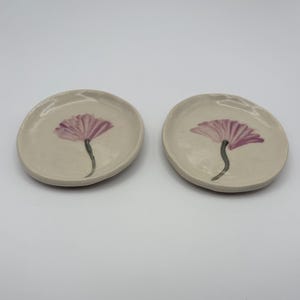 Puede incluir: Dos platos pequeños de cerámica blanquecina. Cada plato presenta una flor rosa pintada a mano con un tallo verde. El diseño floral está centrado en el plato. Los platos son redondos con bordes ligeramente irregulares.