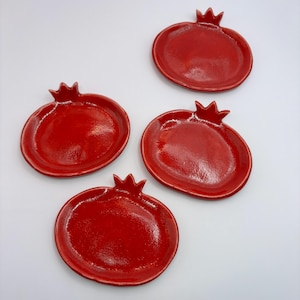 May include: Four glossy red ceramic pomegranate-shaped dishes. Each dish has a raised rim and a crown-like detail at the top. The dishes are arranged on a white surface, showcasing their vibrant colour and unique design.