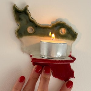 May include: A ceramic candle holder in the shape of a country, with a green and white top and a red base. A lit tea light candle sits on the holder. The holder is being held by a hand with red nail polish.
