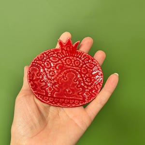 Op de afbeelding: Een glanzend rood keramieken schaaltje in de vorm van een granaatappel, voorzien van een verhoogd, ingewikkeld kantachtig patroon over het oppervlak en een kleine, puntige kroon aan de bovenkant. Het schaaltje heeft een gladde, reflecterende afwerking.