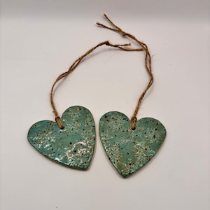 Könnte beinhalten: Zwei türkisfarbene Keramik-Herzornamente mit strukturiertem floralem Design und braunen Bindfäden. Die Herzen haben ein gesprenkeltes Aussehen vor einem weißen Hintergrund. Dekorative Wohnkultur.
