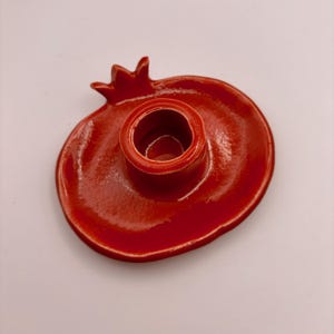 Puede incluir: Un portavelas de cerámica roja con forma de granada. El portavelas tiene una abertura circular central para una vela y una superficie texturizada. El diseño incluye un tallo y una base, todo en un acabado rojo brillante.