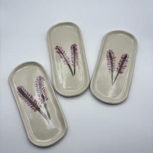 May include: Three off-white ceramic trays with rounded edges. Each tray features a hand-painted design of purple lavender sprigs with green stems. The trays are arranged on a white surface, showcasing a minimalist aesthetic.