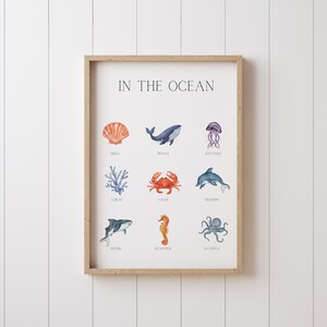 May include: A framed print titled "IN THE OCEAN" featuring watercolor illustrations of marine life. The artwork includes a shell, whale, jellyfish, coral, crab, dolphin, shark, seahorse, and octopus. The frame is a light wood color.