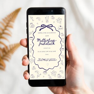May include: A smartphone displays a digital invitation for a Mother's Day brunch. The invitation features a navy blue decorative border with the text "HERZLICHE EINLADUNG ZUM Muttertags-frühstück" and decorative illustrations. The phone is held in a hand.