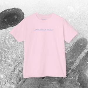 May include: A pink t-shirt with the text "mermaid muse" in a light blue color on the front. The text is in a cursive font.