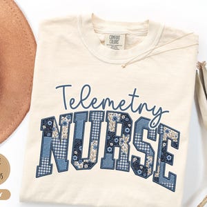 May include: Ivory-colored t-shirt with the words "Telemetry Nurse" in blue script and block letters. The word "NURSE" is designed with a patchwork pattern of blue floral and gingham fabrics. A gold necklace and a brown hat are visible.