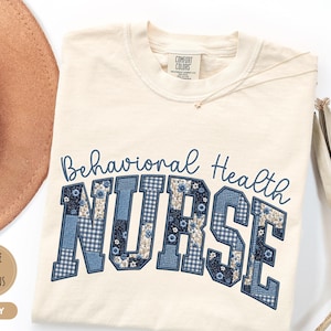 May include: An ivory short-sleeve t-shirt featuring the text Behavioral Health in blue script above the word NURSE in large block letters. The letters are filled with a blue and white patchwork pattern of floral and gingham prints.