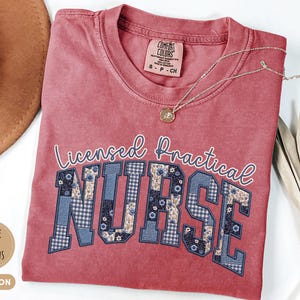 May include: Crimson-colored t-shirt with the words "Licensed Practical Nurse" in white script and block letters. The letters "NURSE" are filled with a blue floral and gingham pattern. A gold necklace with a pendant rests on the shirt.