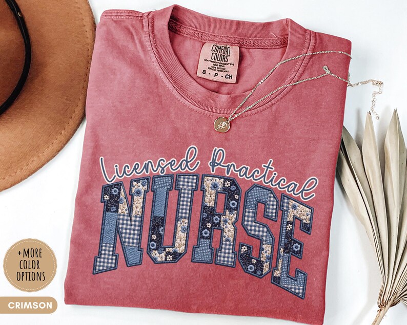 May include: Crimson-colored t-shirt with the words "Licensed Practical Nurse" in white script and block letters. The letters "NURSE" are filled with a blue floral and gingham pattern. A gold necklace with a pendant rests on the shirt.