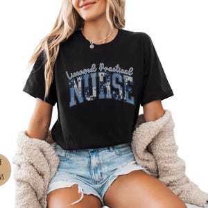 May include: Black t-shirt with the words "Licensed Practical Nurse" in a blue and white patterned design. The shirt is worn with denim shorts and a cream-colored cardigan.