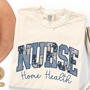 May include: Ivory-colored t-shirt with the word "NURSE" in a patchwork design of blue floral and gingham patterns. Below, the words "Home Health" are embroidered in a matching blue script. A gold necklace and a brown hat are visible.