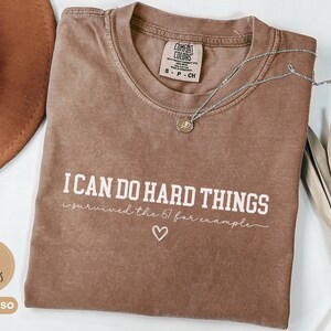 May include: Espresso-colored t-shirt with the text "I CAN DO HARD THINGS" in white. The shirt also features the text "survived the 67 for example" and a heart symbol. A silver necklace with a pendant is draped on the shirt.