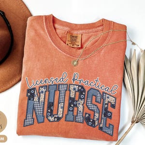 May include: A coral-colored t-shirt with the words "Licensed Practical NURSE" in a collegiate style. The letters are a mix of blue floral and plaid patterns. A gold necklace and a brown hat are also in the image.