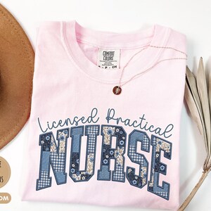 May include: A light pink t-shirt with the words "Licensed Practical Nurse" in a blue, patchwork-style font. The shirt has a crew neck and short sleeves. A gold necklace with a small pendant is draped on the shirt.