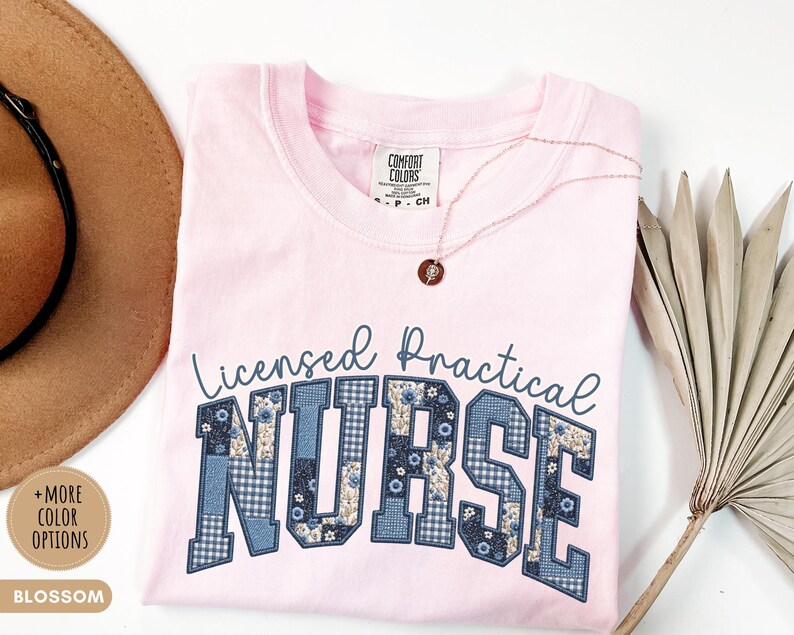 May include: A light pink t-shirt with the words "Licensed Practical Nurse" in a blue, patchwork-style font. The shirt has a crew neck and short sleeves. A gold necklace with a small pendant is draped on the shirt.