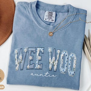 May include: A blue jean colored t-shirt with a crew neckline and short sleeves. The front features the text WEE WOO in large block letters with a blue and white floral and gingham pattern, and the word auntie in white cursive script below.