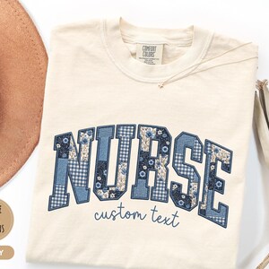 May include: Ivory-colored t-shirt with the word "NURSE" in a blue patchwork design. The design includes floral and gingham patterns. The text "custom text" is below the word "NURSE". A gold necklace and a brown hat are visible.