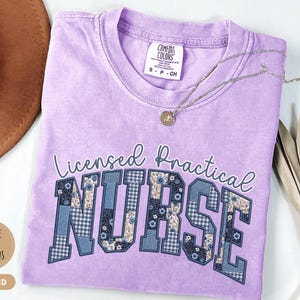 May include: Orchid-colored t-shirt with the words "Licensed Practical Nurse" in a decorative font. The letters "NURSE" are filled with a patchwork design of blue floral and gingham patterns. A silver necklace with a pendant rests on the shirt.