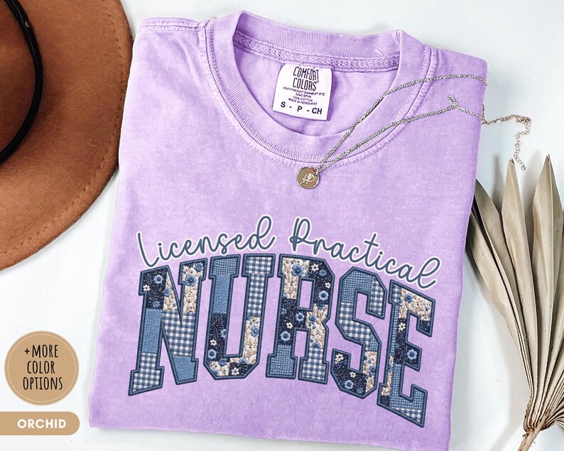 May include: Orchid-colored t-shirt with the words "Licensed Practical Nurse" in a decorative font. The letters "NURSE" are filled with a patchwork design of blue floral and gingham patterns. A silver necklace with a pendant rests on the shirt.