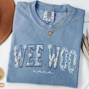 May include: A blue jean colored t-shirt with a crew neckline and a Comfort Colors brand label. The shirt features the text WEE WOO in large block letters with a blue and white floral and gingham pattern, and the word nana in white script below.