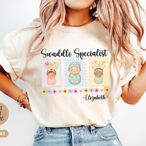 May include: Ivory-colored t-shirt with the text "Swaddle Specialist" above three postage stamp designs featuring swaddled babies. The name "Elizabeth" is printed below the stamps. The shirt also has a "+MORE COLOR OPTIONS" button.