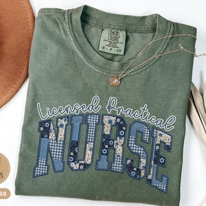 May include: Moss green t-shirt with the words "Licensed Practical Nurse" in white script and block letters. The letters "NURSE" are decorated with blue floral and gingham patterns. A gold necklace with a pendant rests on the shirt.