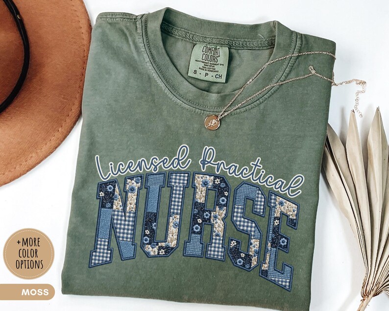 May include: Moss green t-shirt with the words "Licensed Practical Nurse" in white script and block letters. The letters "NURSE" are decorated with blue floral and gingham patterns. A gold necklace with a pendant rests on the shirt.