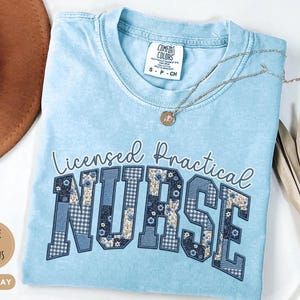 May include: Light blue t-shirt with the words "Licensed Practical Nurse" in a decorative font. The word "NURSE" is in a patchwork design with floral and gingham patterns. A gold necklace and a brown hat are also in the image.