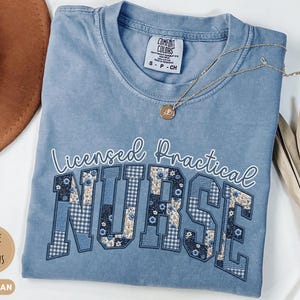 May include: Blue jean colored t-shirt with the words "Licensed Practical Nurse" in white script and patchwork letters. The "NURSE" letters feature floral and gingham patterns. A gold necklace with a pendant rests on the shirt.