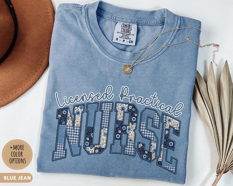 May include: Blue jean colored t-shirt with the words "Licensed Practical Nurse" in white script and patchwork letters. The "NURSE" letters feature floral and gingham patterns. A gold necklace with a pendant rests on the shirt.