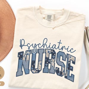 May include: Ivory-colored t-shirt with the words "Psychiatric Nurse" in blue script and patchwork letters. The letters are decorated with floral and gingham patterns. A gold necklace and a brown hat are visible.