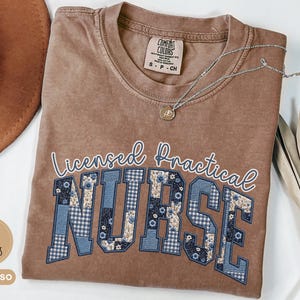 May include: Espresso-colored t-shirt with the words "Licensed Practical Nurse" in white script and blue patchwork letters. A silver necklace with a small pendant rests on the shirt. The shirt has a Comfort Colors tag.