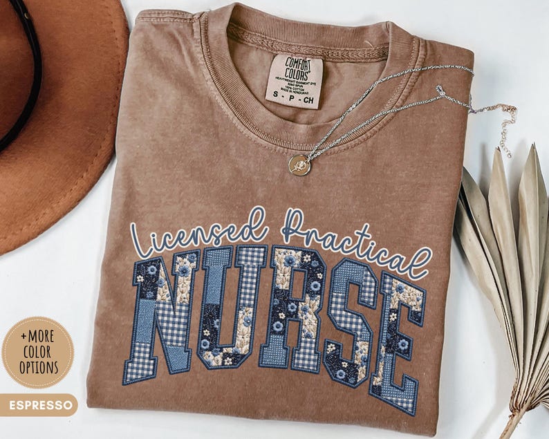 May include: Espresso-colored t-shirt with the words "Licensed Practical Nurse" in white script and blue patchwork letters. A silver necklace with a small pendant rests on the shirt. The shirt has a Comfort Colors tag.